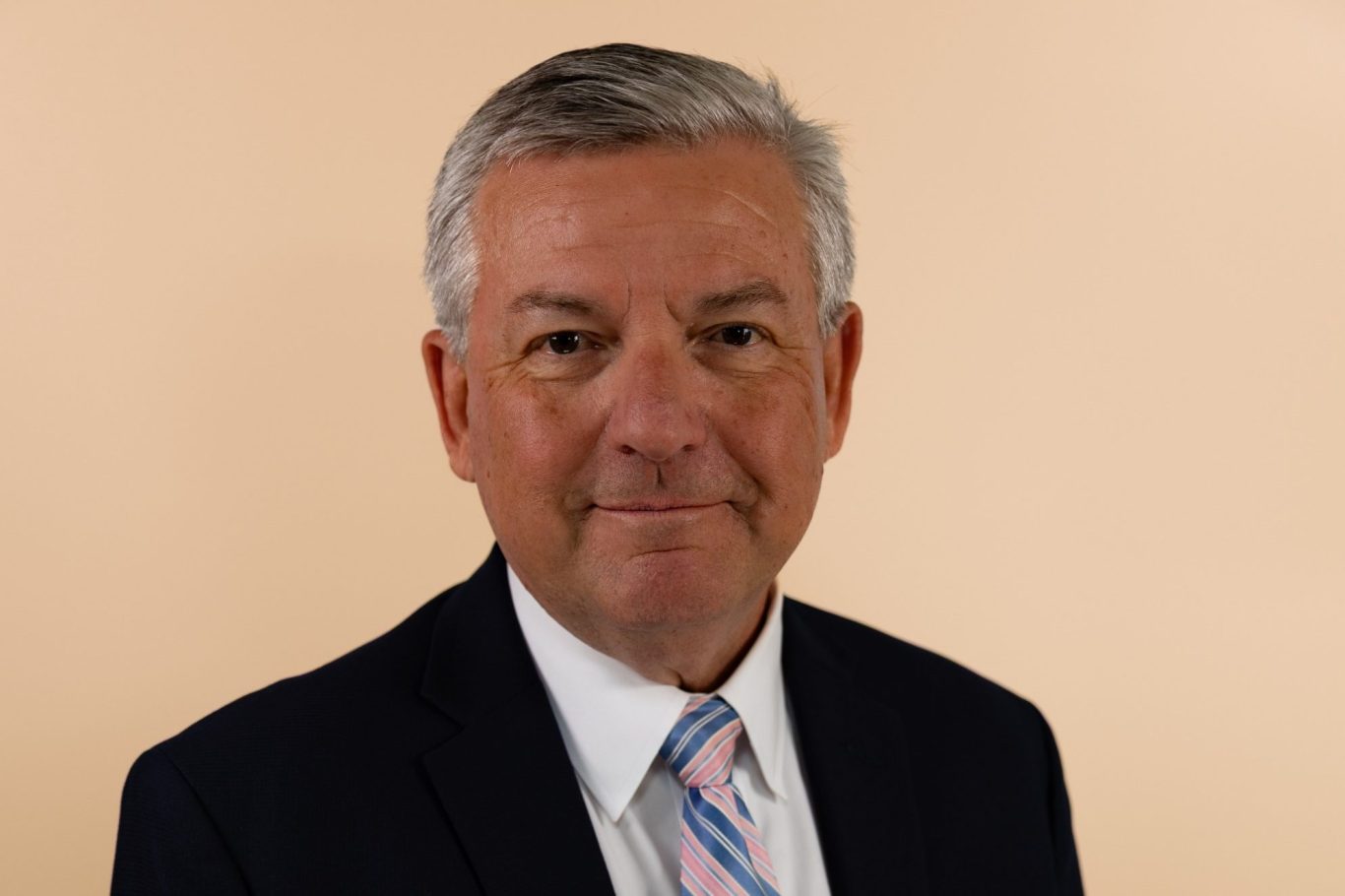 Corporate headshot Man in a dark suit with a light pink tie, smiling against a soft beige background.