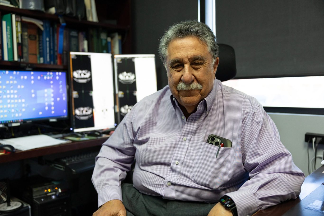 Testimonial and business interviews A man with a mustache sits in an office, surrounded by books and two printed images.