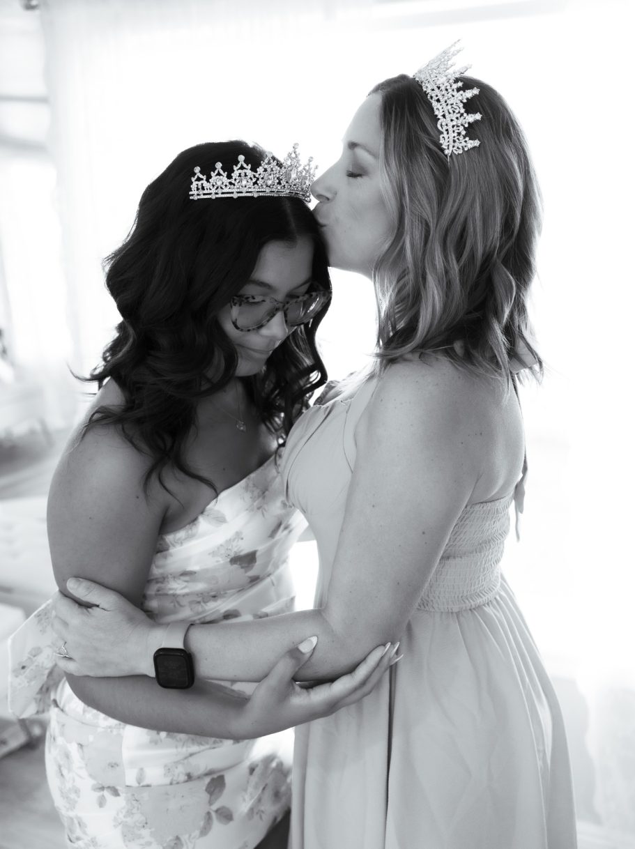 Two women, both wearing tiaras, embrace affectionately in a softly lit setting.