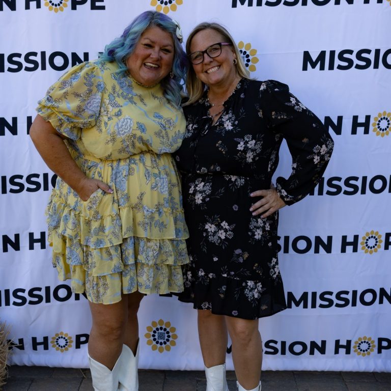 Mission Hope Two women smiling together, one in a yellow dress and the other in a black floral dress.