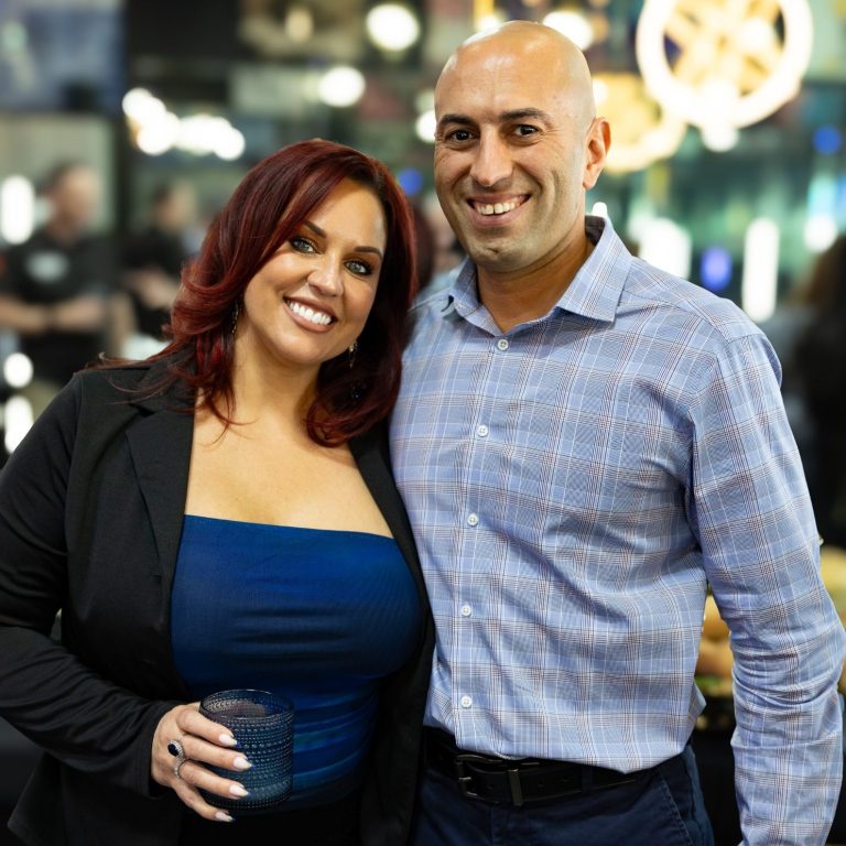 Blue Vibes Hair Studio Smiling man and woman pose together at a social event, with a drink in the woman's hand.