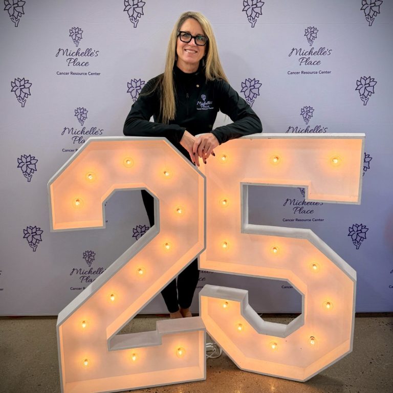 Michelle's Place Cancer Resource Center Woman in glasses poses beside large illuminated numbers "25" at a celebration event.